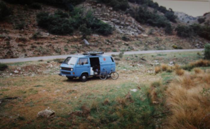 Blue T25 panel van parked in a field beside a mountain road
