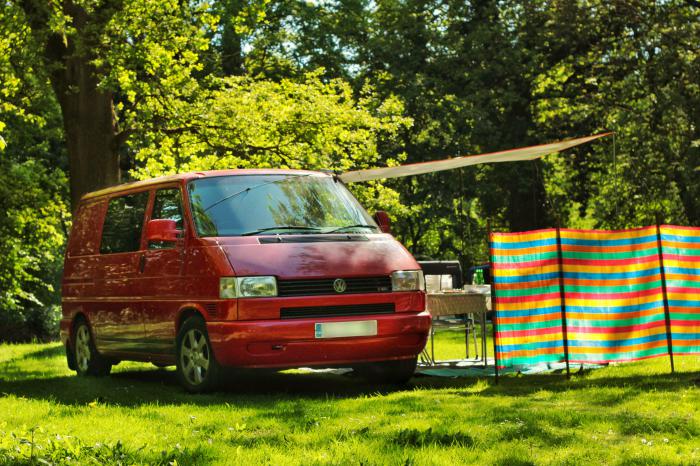 Red VW T4 parked in a sunny field beside a striped windbreak